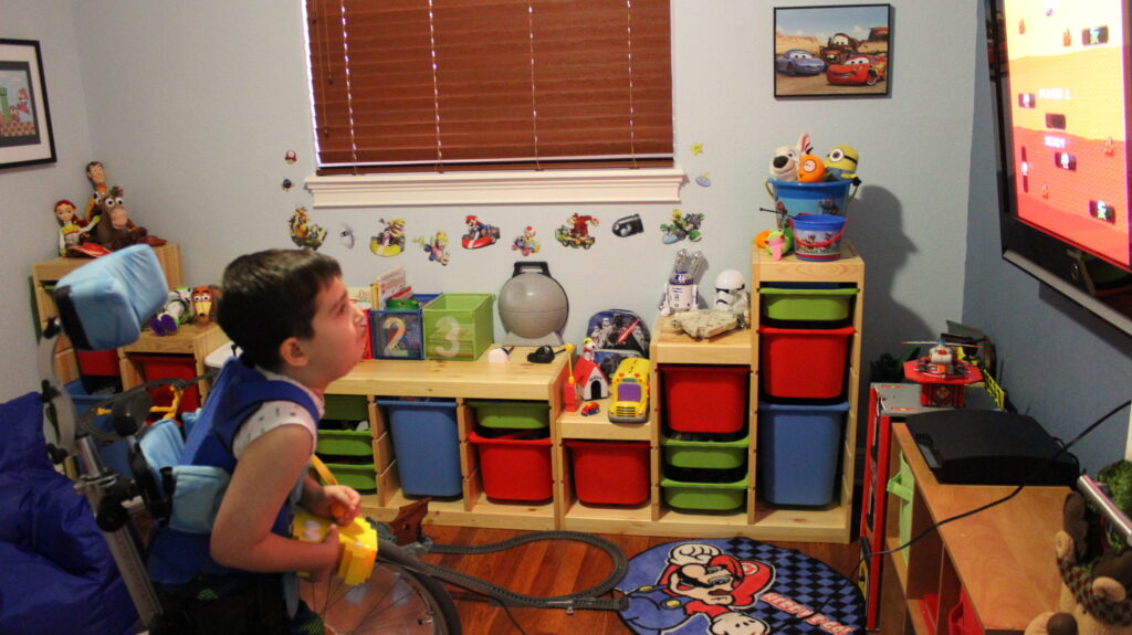 A young boy in a supportive wheelchair in his playroom, facing shelves of toys