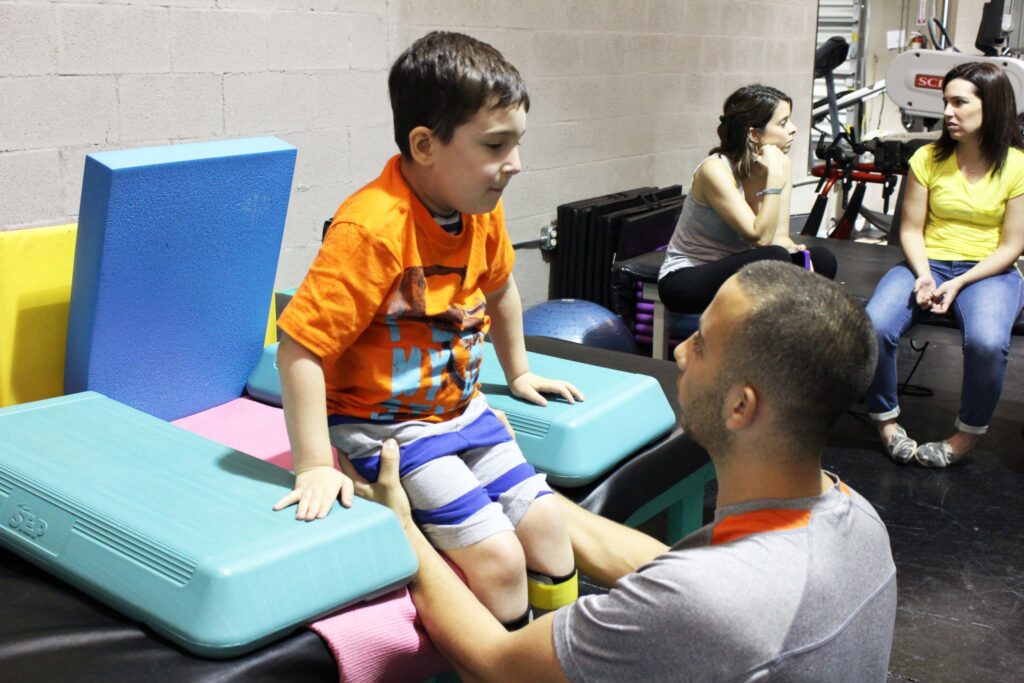 A young boy sitting on a therapy block while a man talks to him, two women watching