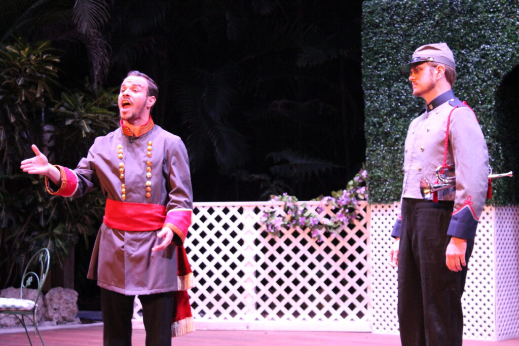 Two actors in grey uniforms with red sashes talking on stage in front of a white lattice