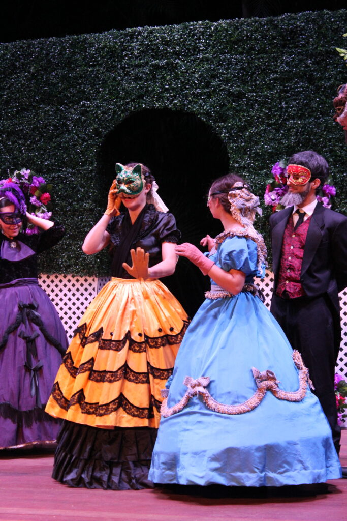 Three actors in masks and hoop-skirted gowns on stage in front of a green hedge