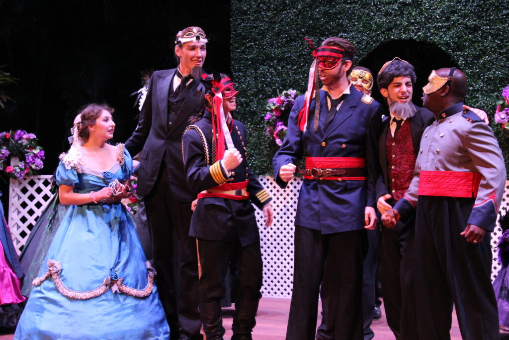 Actors in period uniforms with red sashes and gowns on stage, some in masks