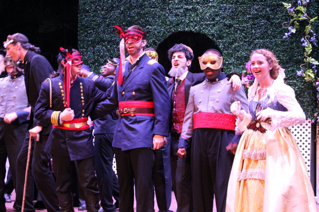 A cast in period uniforms and masks gathered on stage, a woman in a white gown among them
