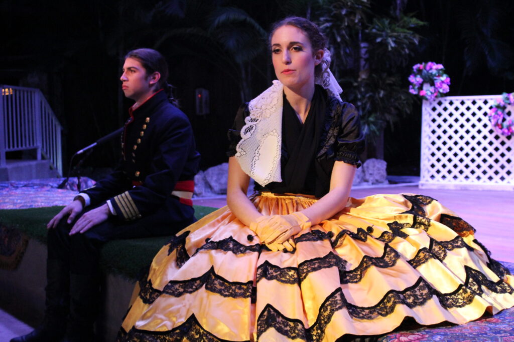 An actress in a yellow-and-black hoop skirt sitting on stage beside an actor in uniform