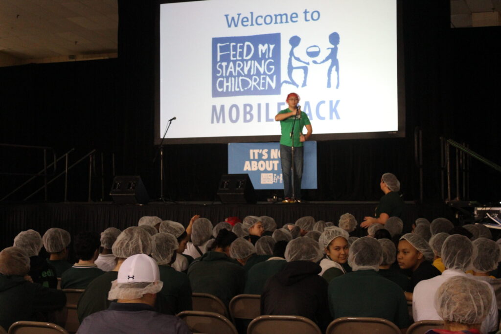 A hall full of volunteers in hairnets facing a stage and a screen reading "Welcome to Feed My Starving Children MobilePack"