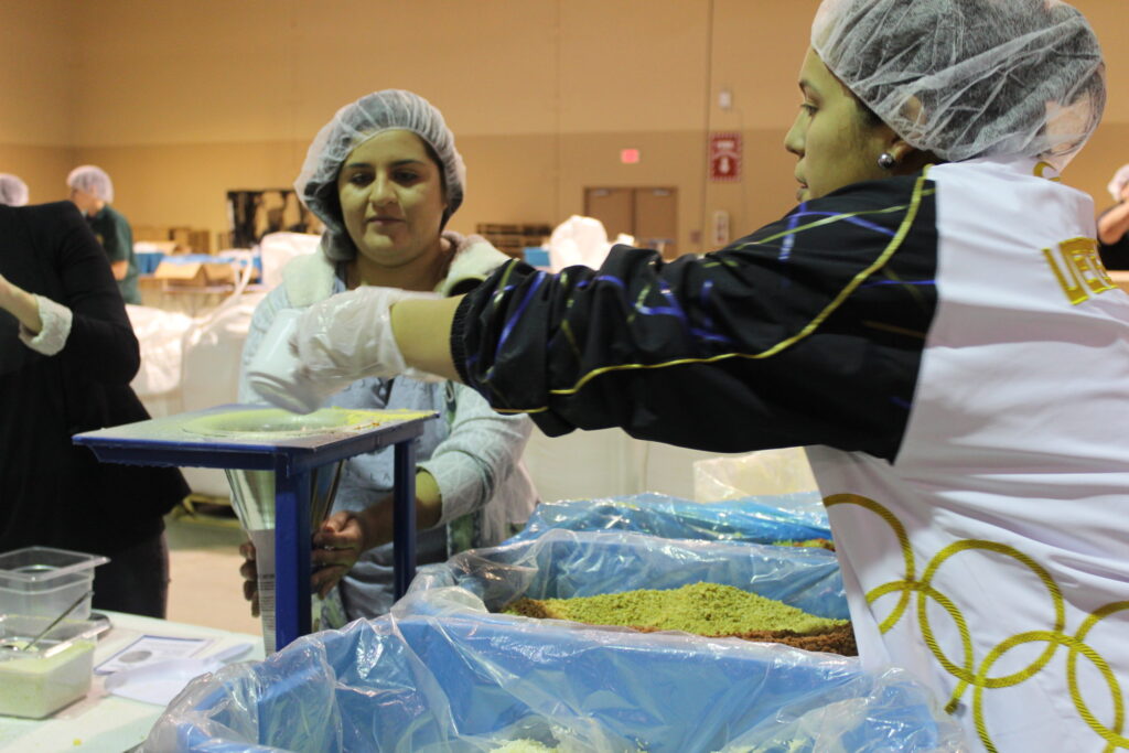 Volunteers in hairnets and gloves pouring ingredients into bags at a packing station