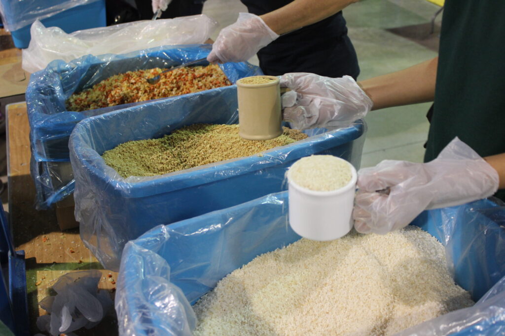 Gloved hands measuring rice and dried vegetables from blue-lined bins into meal packs