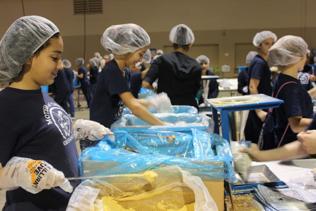 Young volunteers in hairnets filling meal bags at long tables in a hall