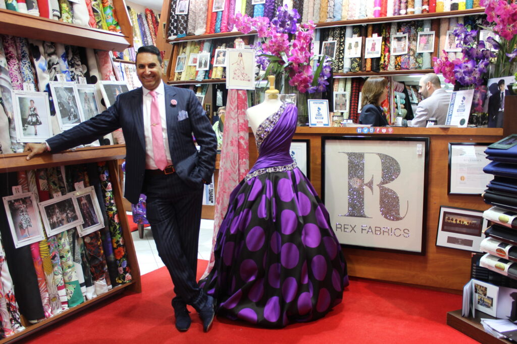 A man in a suit and pink tie standing beside a purple polka-dot ball gown in a fabric shop