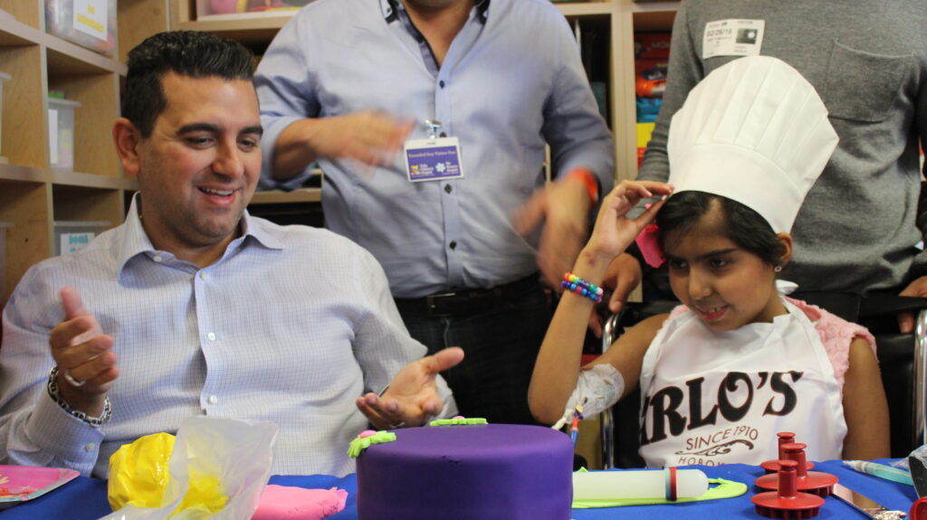Buddy Valastro laughing beside a girl in a chef's hat at a decorating table