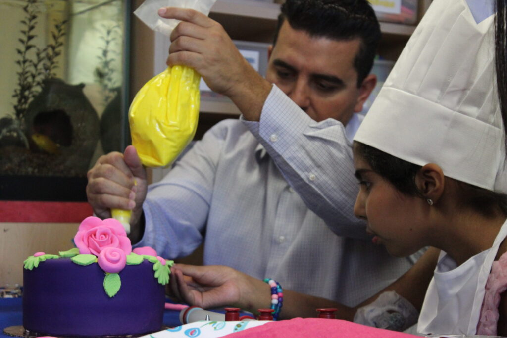 Buddy Valastro helping a girl in a chef's hat pipe yellow icing onto a purple cake