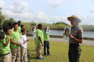 A park ranger in a hat showing a card to fourth-graders in green T-shirts beside a lake