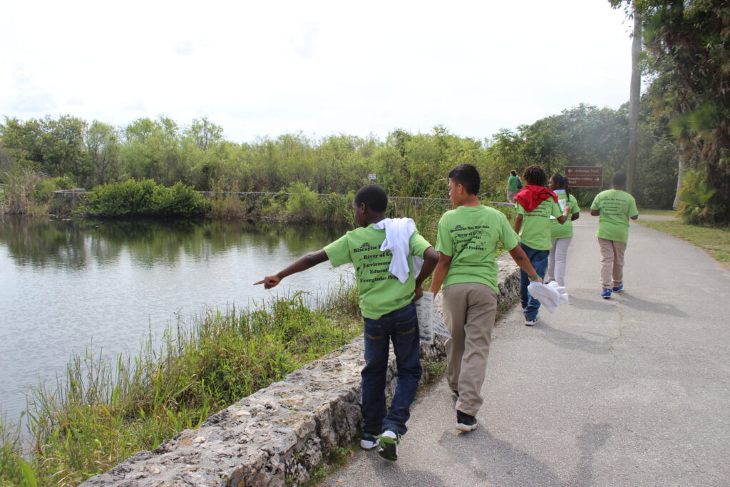 Children in green T-shirts walking along a path beside a lake, one pointing