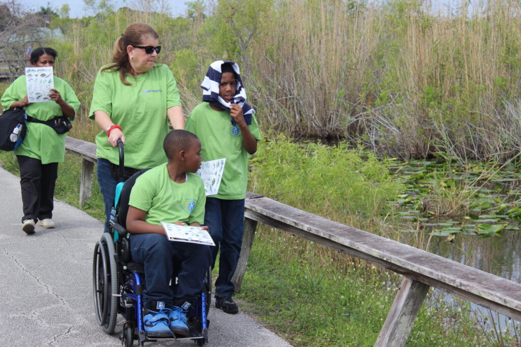 Children and adults in green T-shirts on a boardwalk, one boy in a wheelchair
