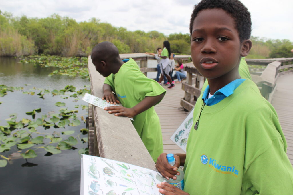Boys in green Kiwanis T-shirts on a boardwalk over a lily-covered pond, one holding a worksheet