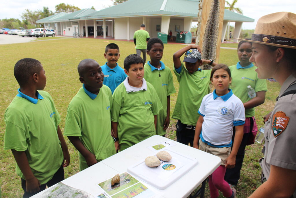 A park ranger talking to schoolchildren in green T-shirts at an outdoor table of exhibits