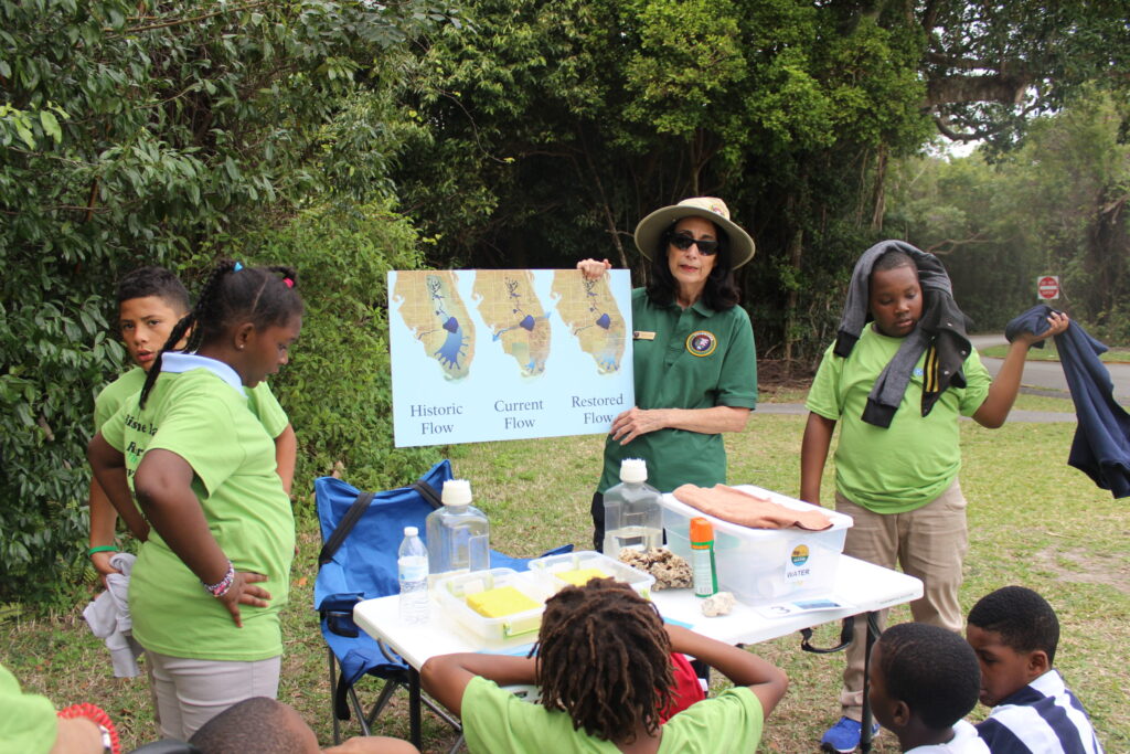 A ranger showing a map of Florida to children in green T-shirts under the trees