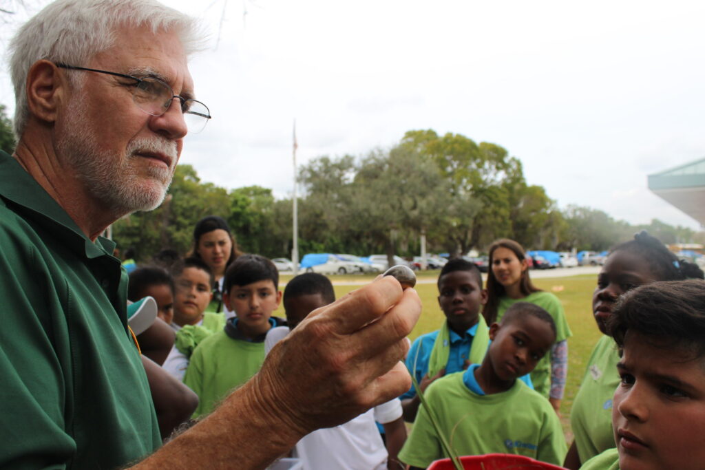 An older man in glasses holding up something small for a group of watching children