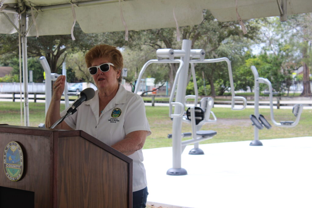 A woman in white sunglasses speaking at a lectern in front of new outdoor fitness equipment