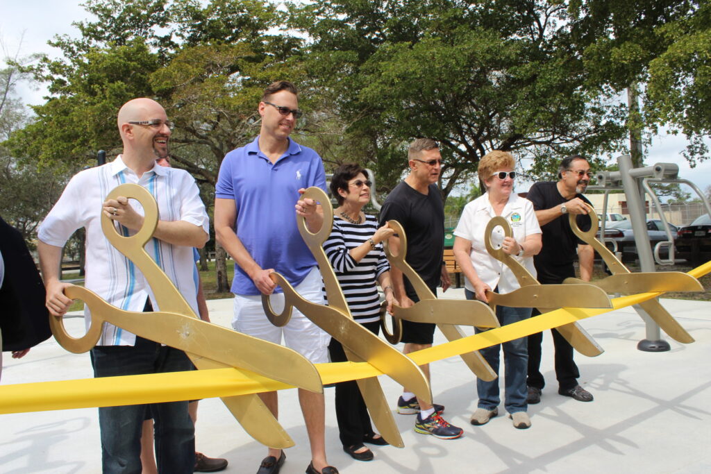 A row of people holding giant gold scissors to cut a yellow ribbon at a park opening