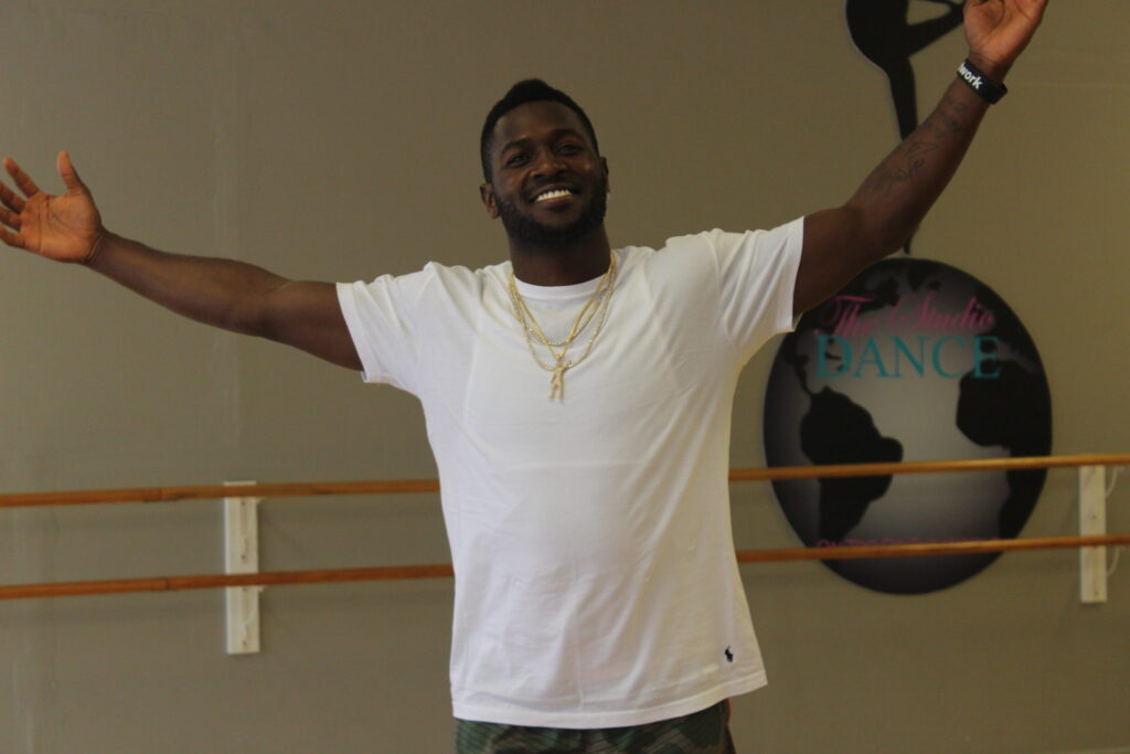 Antonio Brown in a white T-shirt and gold chain, arms spread wide in a dance studio