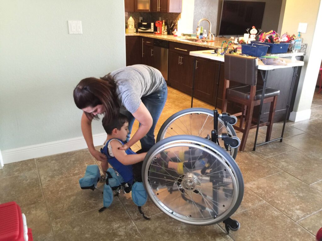 A woman lifting a young boy into his wheelchair in a kitchen