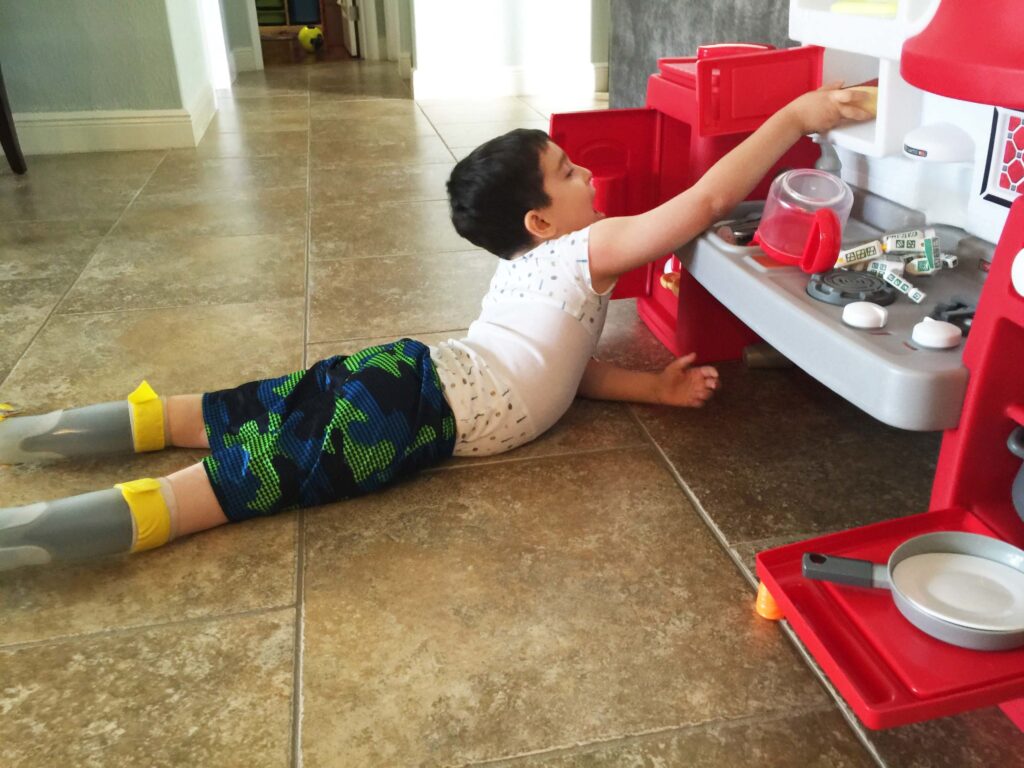 A young boy lying on the floor reaching up to a red toy kitchen