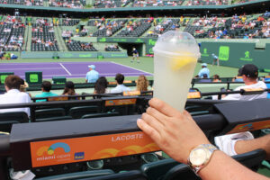 A hand holding a lemonade in a plastic cup over the stands at a Miami Open tennis match