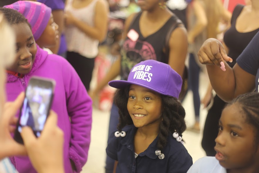 A girl in a purple cap smiling as another child films her on a phone