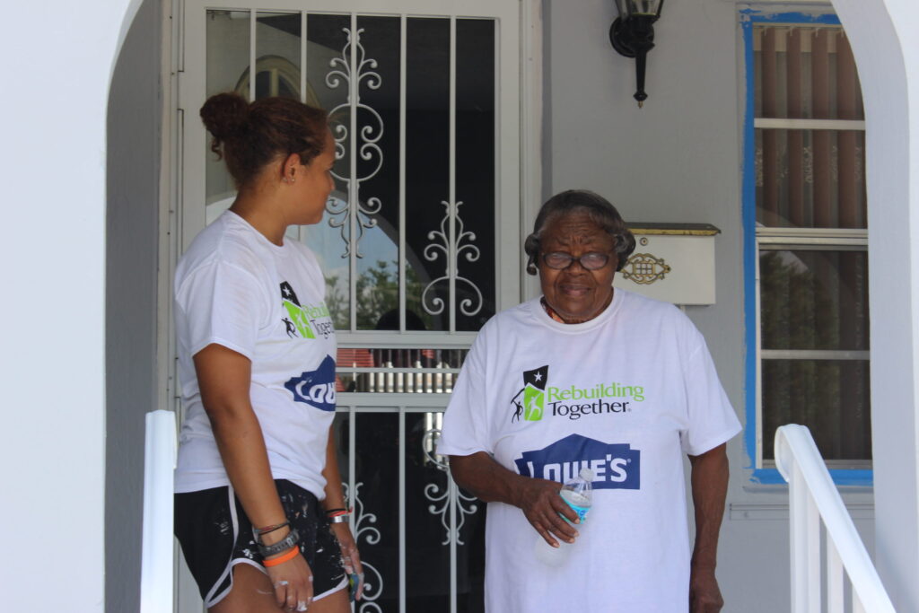 A volunteer in a Rebuilding Together T-shirt standing with an elderly woman at her front door