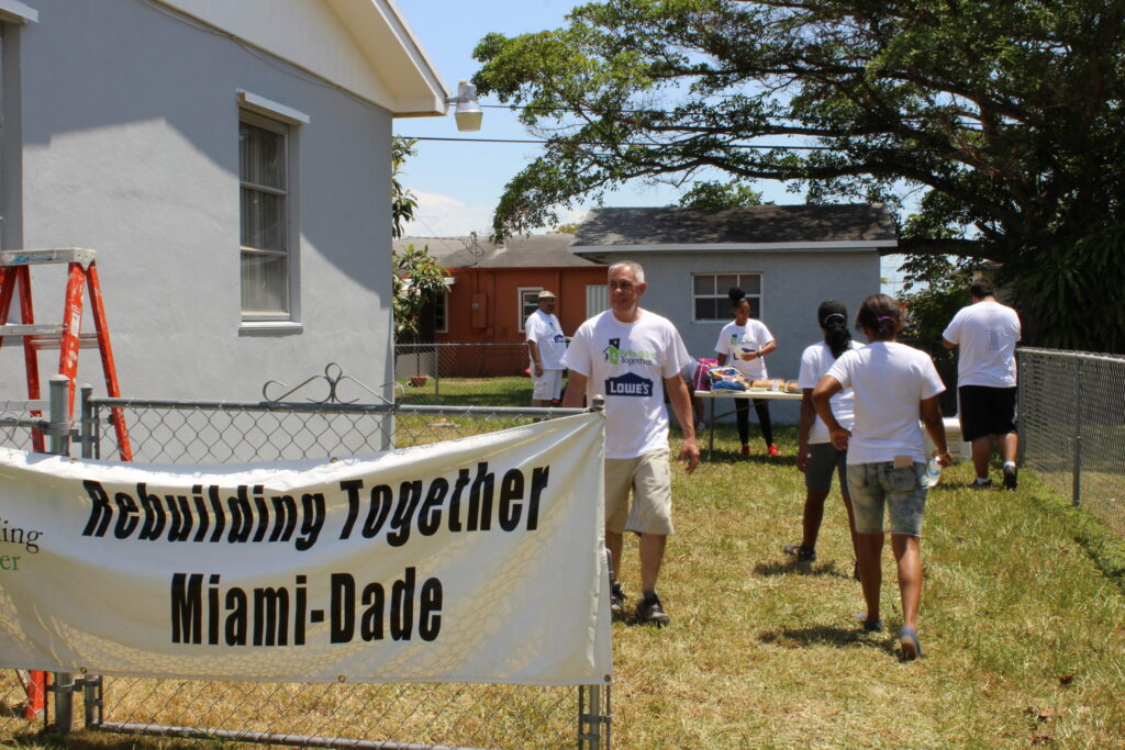 Volunteers working on a house behind a "Rebuilding Together Miami-Dade" banner on a fence