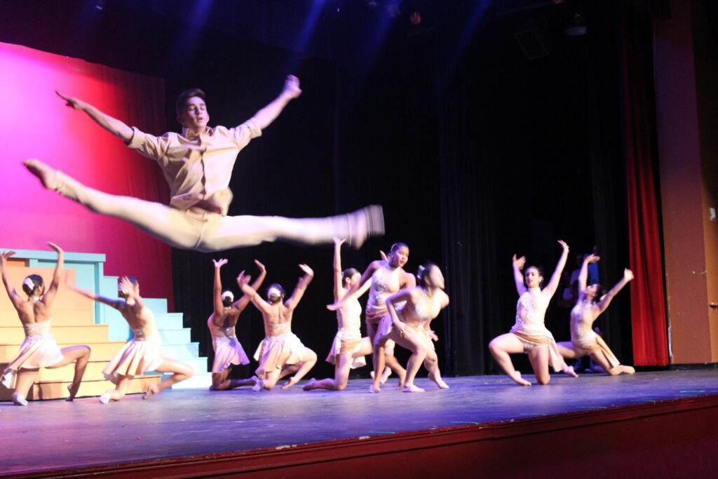 A student dancer in a split leap above a line of dancers on a pink-lit school stage