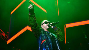 J Balvin in sunglasses and a sequinned jacket, singing and pointing on a green-lit stage