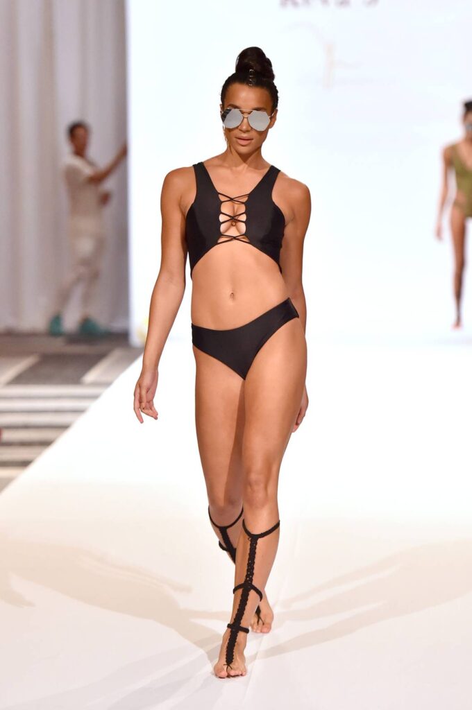 A model in a black strappy swimsuit and lace-up sandals on a white runway