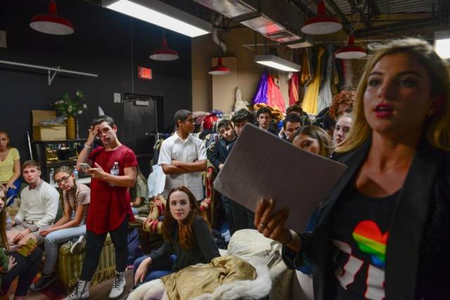A student with a clipboard talking to a room of seated students at rehearsal