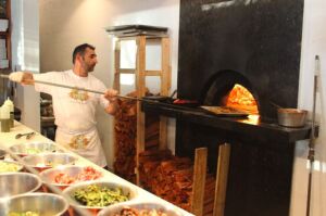 A pizza maker sliding a pizza into a wood-fired oven, bowls of toppings in front of him
