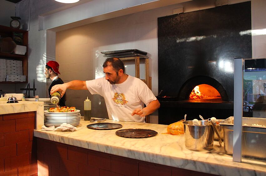 A pizza maker topping pizzas at the counter beside a glowing wood-fired oven