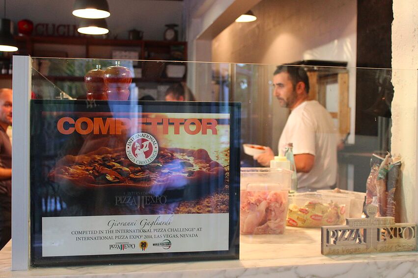 A framed pizza competition certificate on the counter, the pizza maker working behind it