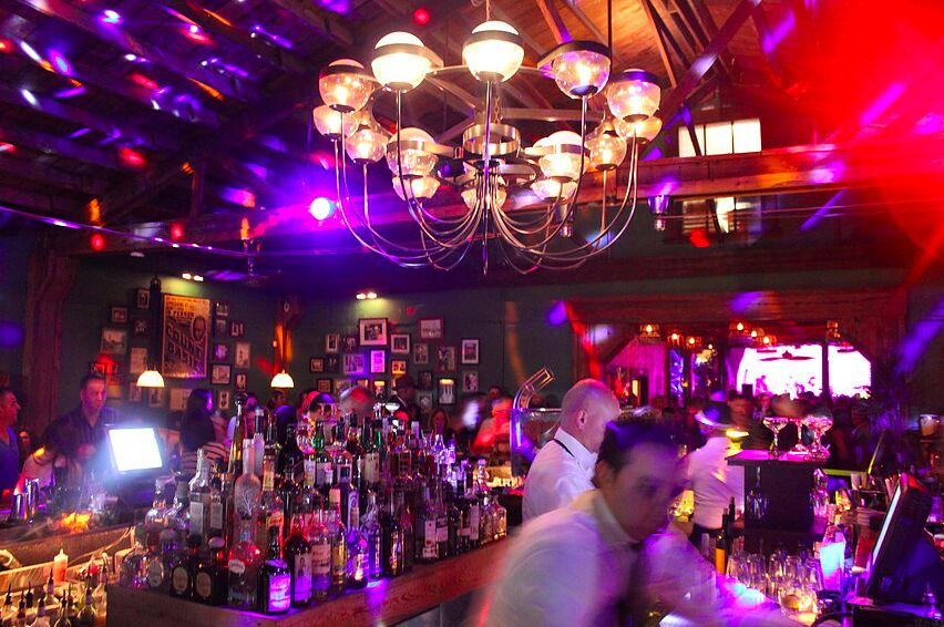 A busy bar lit purple and red under a chandelier, bartenders at work