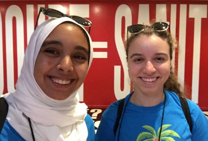 Two young women smiling for a selfie, one in a white hijab, in front of a red and white mural