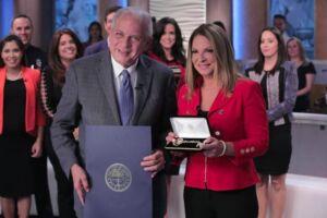 Ana María Polo in a red jacket receiving a key to the city from a man in a suit, colleagues around them