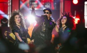 Daddy Yankee in a cap and sunglasses performing with two dancers on a purple-lit stage