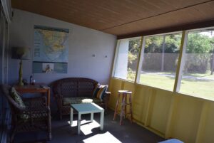 A sunny screened porch with a desk, wicker sofa and a map on the wall, looking out on grass and trees