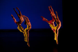 Two dancers in flowing orange costumes leaping in the air against a deep blue backdrop