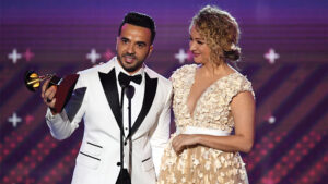 Luis Fonsi in a white tuxedo jacket holding a Latin Grammy at a microphone beside a woman in a cream dress