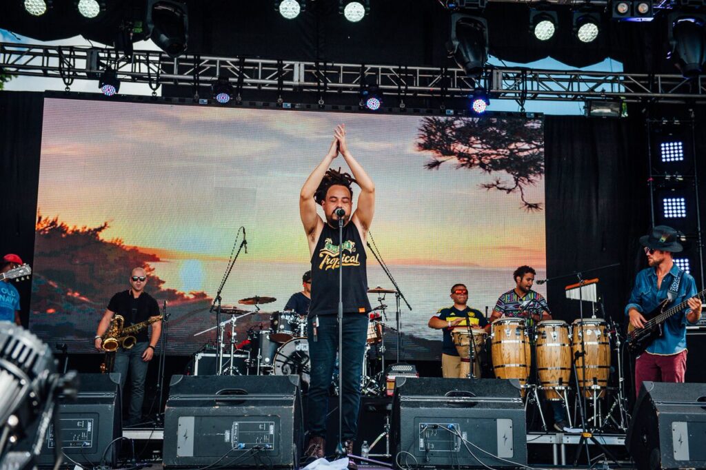 A singer with arms raised in prayer on a festival stage at sunset, a band around him