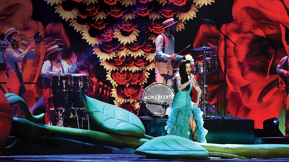 A performer in a green gown on a stage decorated with giant red flowers and leaves, a band behind her