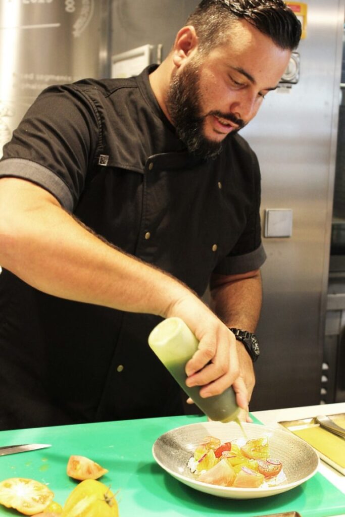 A chef in black squeezing sauce onto a plate in a restaurant kitchen