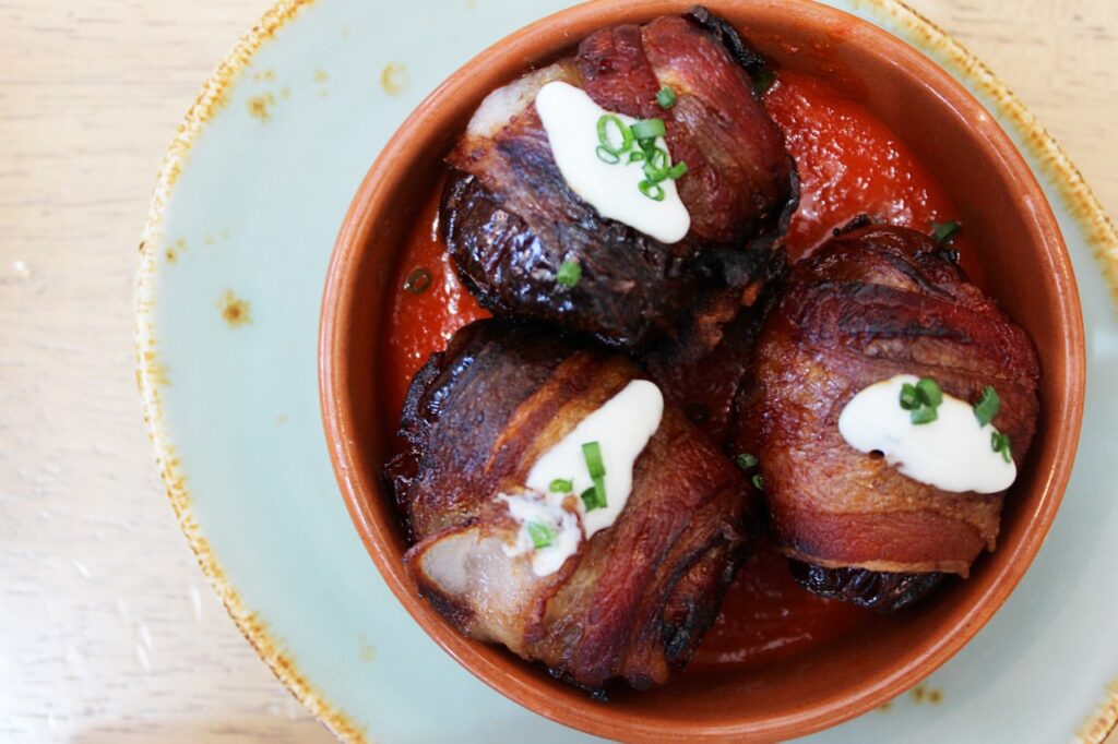 Bacon-wrapped dates in tomato sauce topped with dollops of cream, in a terracotta dish