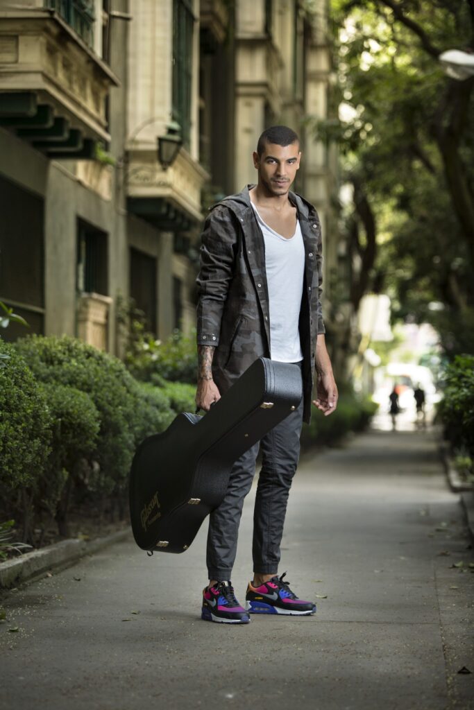 Manuel Medrano standing in a leafy lane holding a guitar case