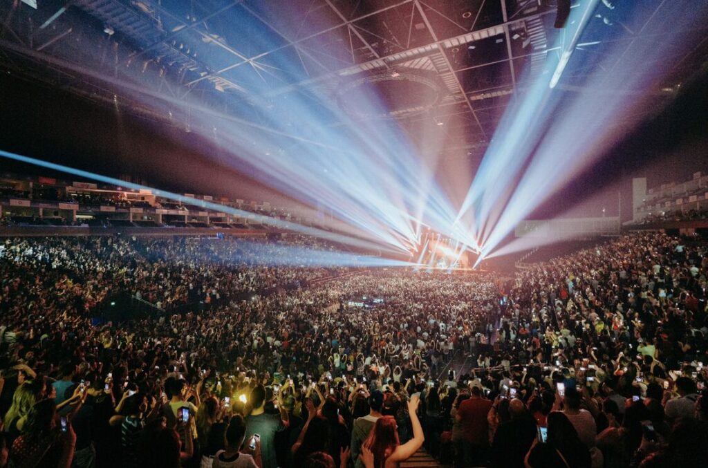 Beams of stage light over a packed arena crowd, seen from the back